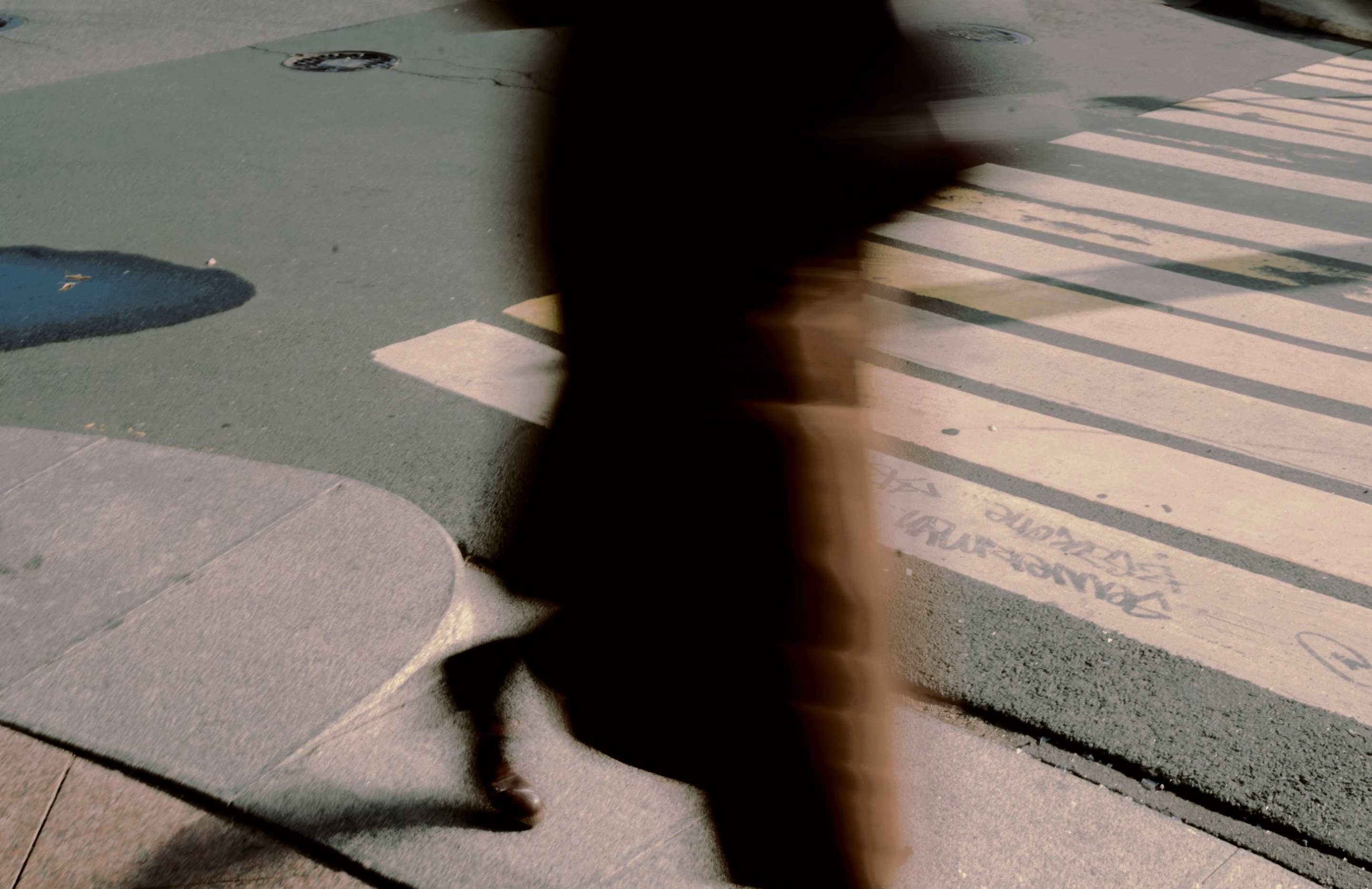 A person in a long coat striding across a sunlit city crossing, blurred by movement