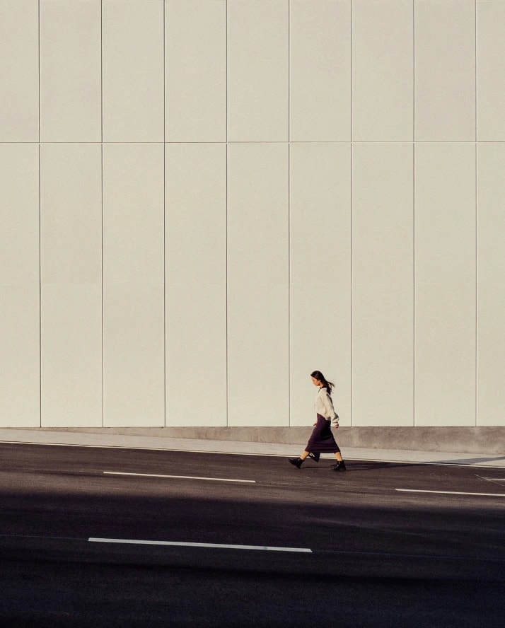 A woman walking past a tall pale wall on an empty street