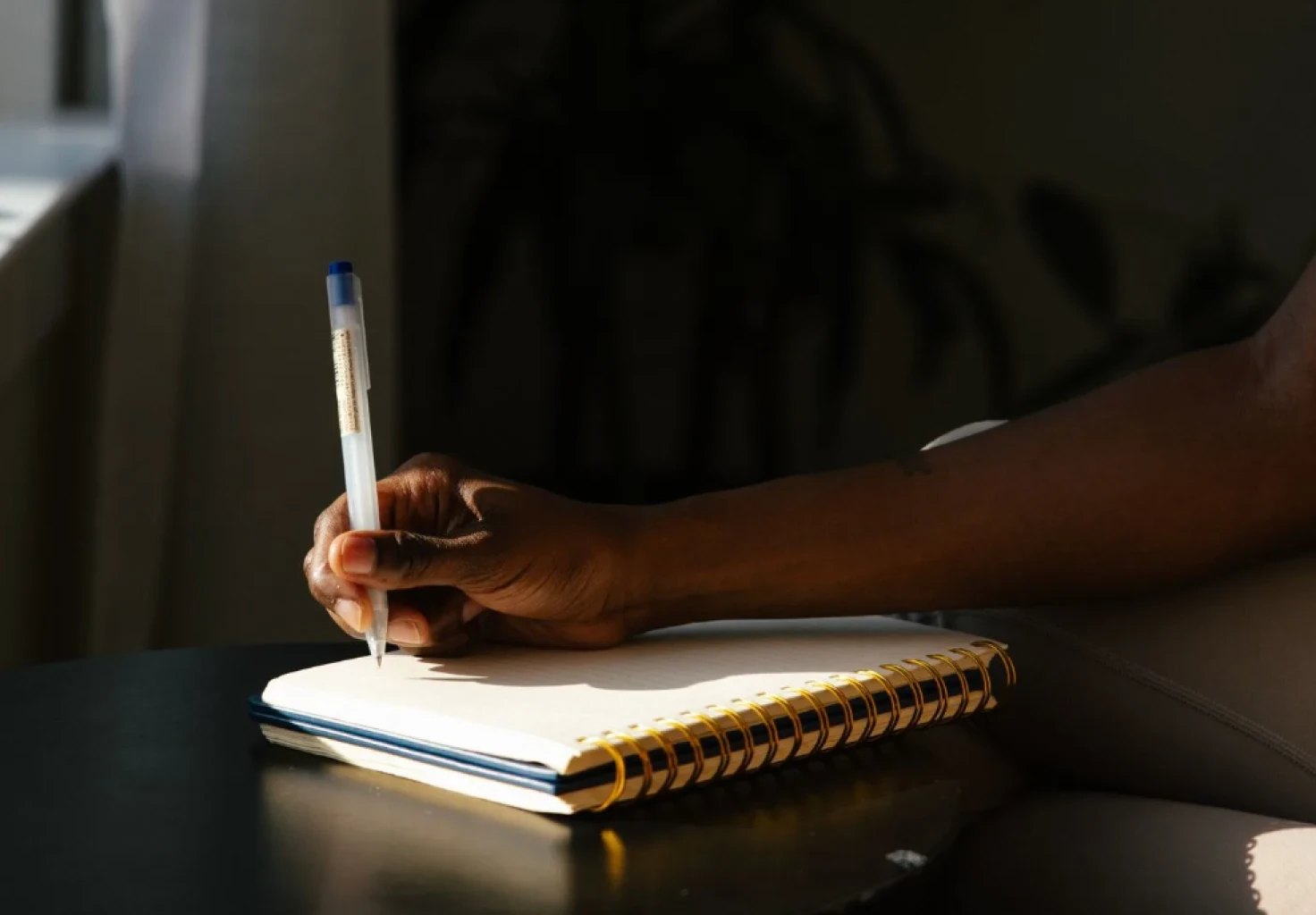 A hand writing in a spiral notebook with a pen, lit by low sun